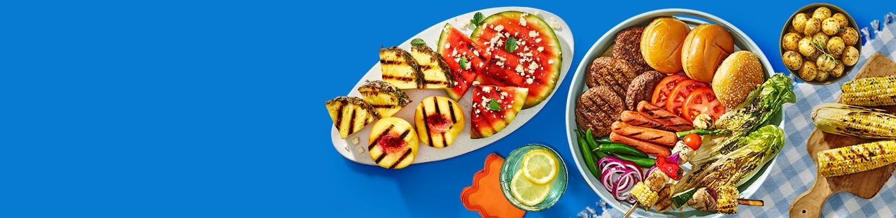 Summer themed table with a checkered picnic blanket sitting underneath a platter of grilling essentials like buns, tomatoes, burgers, hot dogs and lettuce, grilled pineapple and watermelon on a white oval plate, grilled corn on the cob and mini potatoes. 