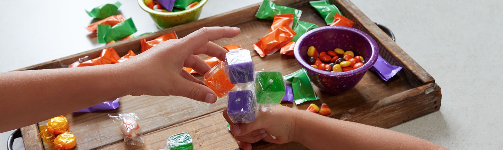 A rustic wooden tray covered in various candy in the background with a childs hand gripping lollipops and an adults hand reaching for them.