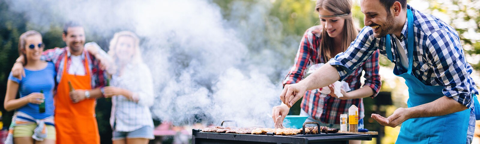 Outdoor grill with two people standing over it turning the hot dogs. In the background a group of friends watches on.