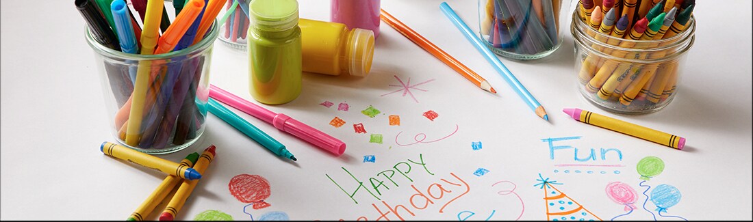 On a paper table cloth sits markers, crayons and other craft supplies with Happy Birthday written on the paper in the middle On a paper table cloth sits markers, crayons and other craft supplies with Happy Birthday written on the paper in the middle
