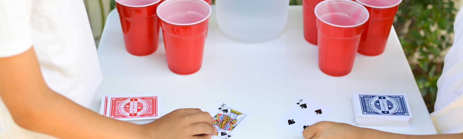 On an outdoor table 2 sets of 4 red solo cups sit with a jug of water in the middle while 2 people stand on each side facing off in a card game.